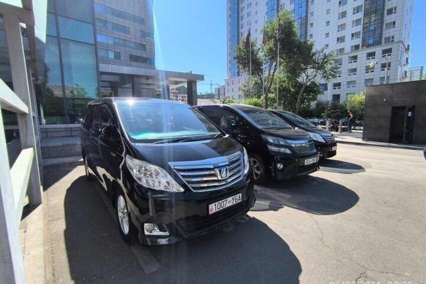 A black van parked in a lot with other vehicles, modern buildings, and a clear sky in the background. 2