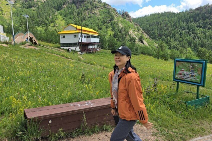 A person walking on a grassy path with a wooden bench, a house with a yellow roof, and a signboard in a hilly landscape. 3