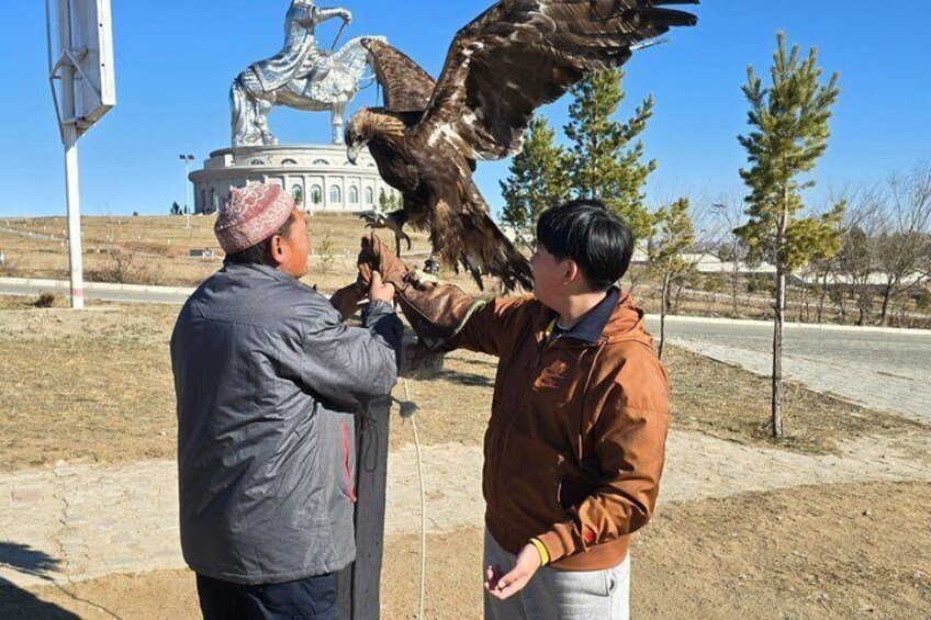 Two people interacting with a large bird of prey, possibly a falcon, in an outdoor setting with a statue and a building in the background. 4
