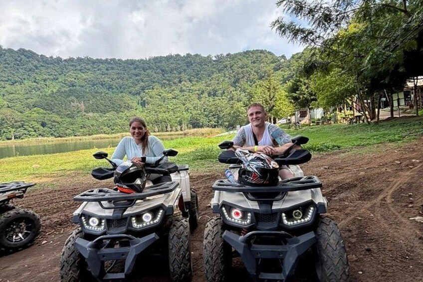 Two people on all-terrain vehicles in a rural setting with a lake and forested hills in the background. 5