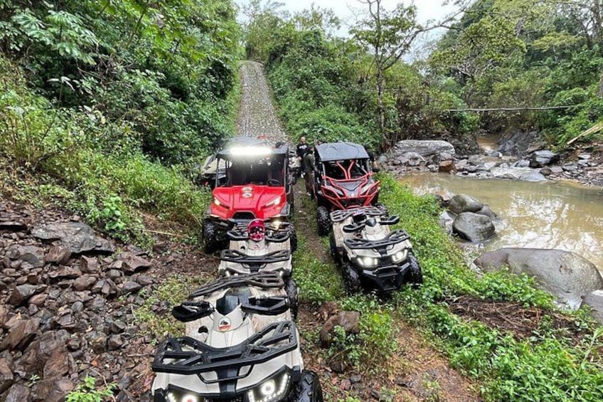 Off-road vehicles parked by a rocky stream in a forested area. 1