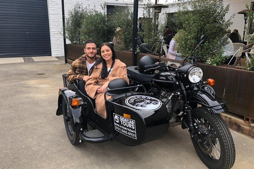 Een man en vrouw zitten op een motor met zijspan, geparkeerd in een open gebied met een gebouw en begroeiing op de achtergrond. 3