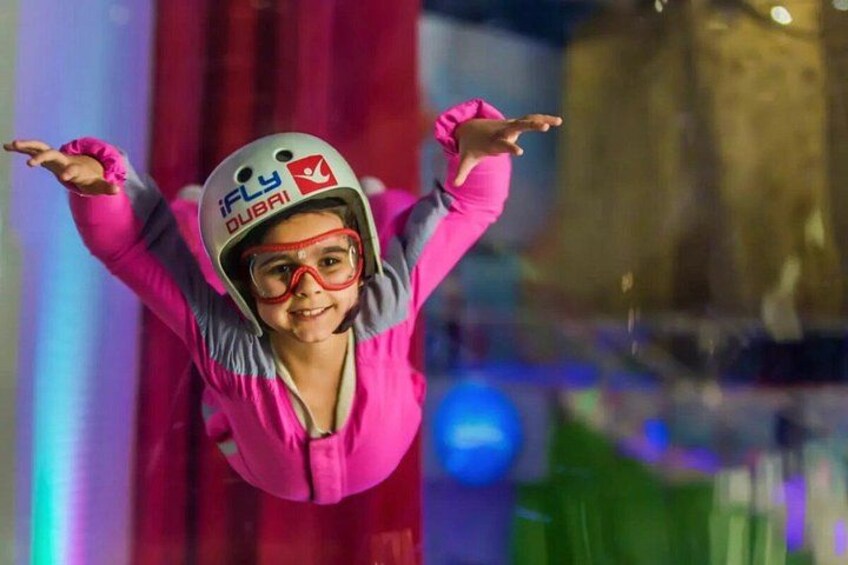 Une personne vêtue d’une tenue rose et portant un casque, les bras tendus, se trouve dans un espace intérieur éclairé de couleurs vives. 1