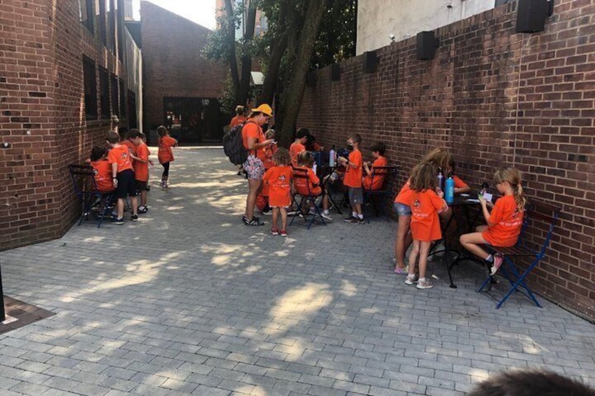 Niños con camisetas anaranjadas reunidos en un espacio al aire libre con paredes de ladrillo y árboles. 1