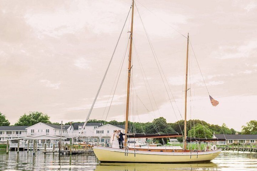 A sailboat with two masts and an American flag, docked near a waterfront with houses in the background. 3