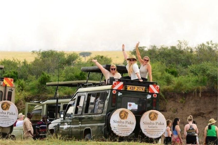 Un véhicule de safari avec des personnes assises sur le toit, entouré d’herbes sèches et de quelques arbres clairsemés. 1