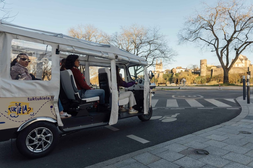 A tuk-tuk with passengers, parked on a city street with trees and buildings in the background. 2