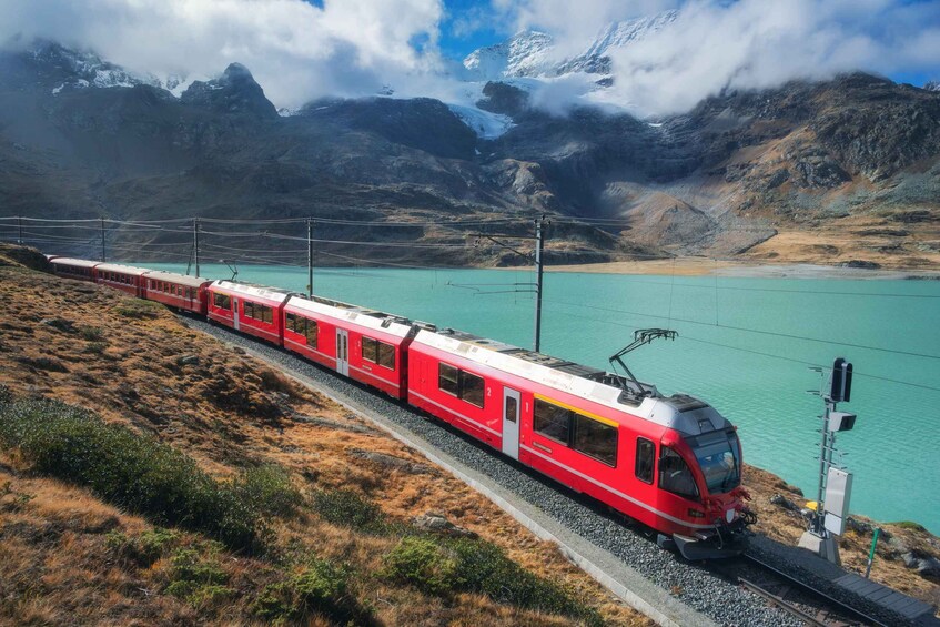 A red train on tracks with a mountainous backdrop and a turquoise lake. 1