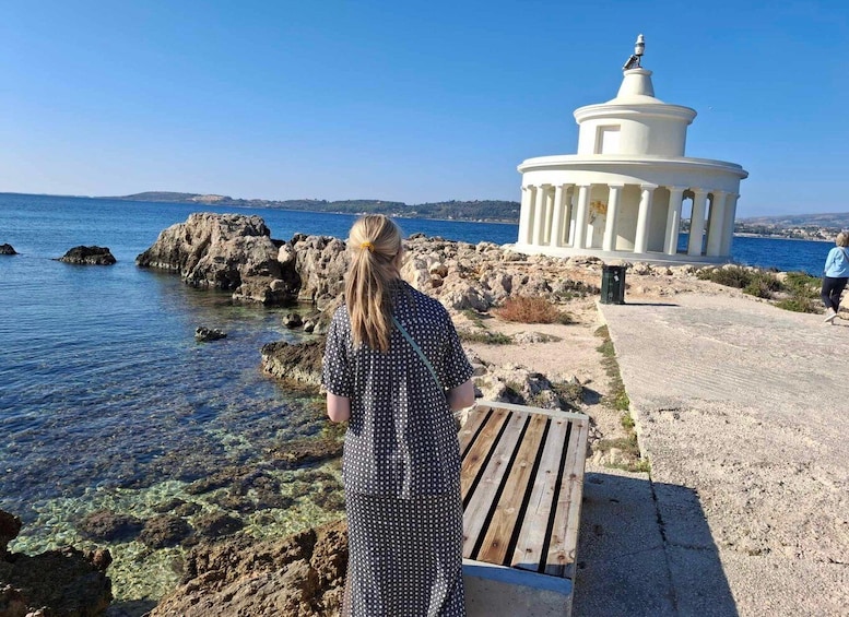 A woman standing by a rocky shoreline with clear water, a white gazebo, and a distant view of the sea. 1