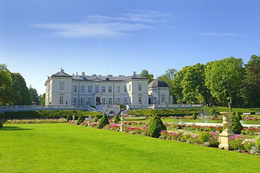 A large, white, classical-style building with a symmetrical facade, surrounded by well-manicured gardens and a clear blue sky. 1