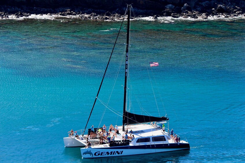 A Gemini sailboat with people on deck, anchored near a rocky shoreline. 5