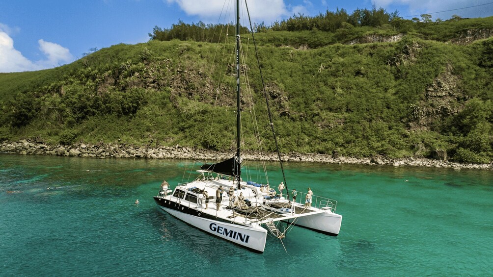 A sailboat named 'GEMINI' anchored in turquoise waters with a forested hillside in the background. 2