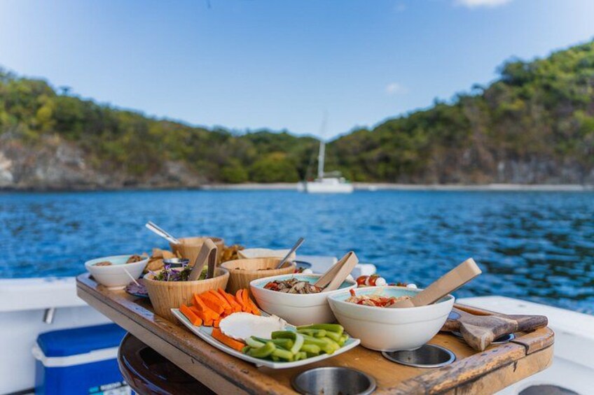 A wooden table with various food items and utensils on a boat, with a body of water and a sailboat in the background. 5