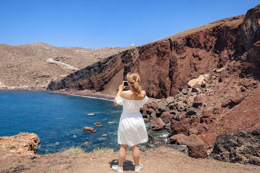 A woman in a white dress taking a photo of a coastal landscape with rocky cliffs and clear blue water. 4