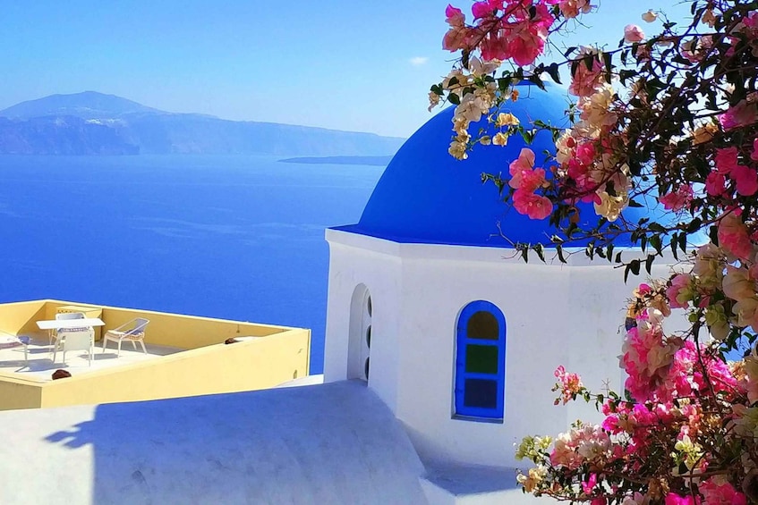 A white building with a blue dome, a yellow terrace with white chairs, and pink flowers. 5