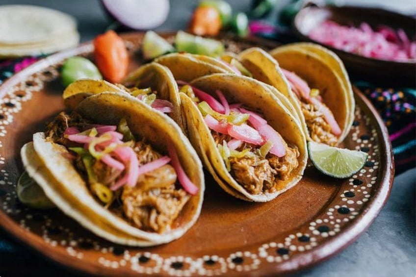 Three tacos with shredded meat, pickled onions, and a lime wedge on a decorative plate. 2