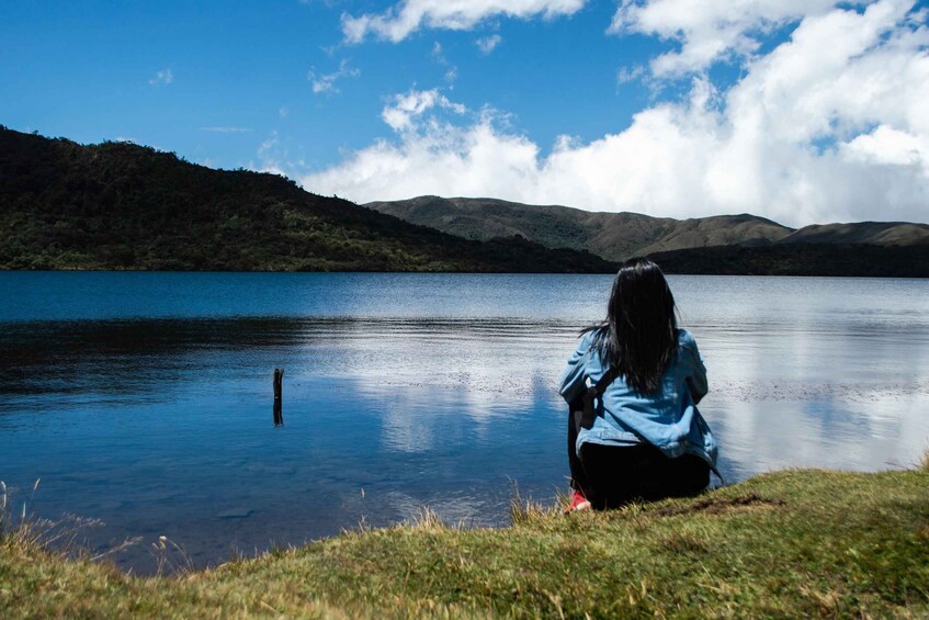 A person sitting by a calm lake with mountains in the background. 1