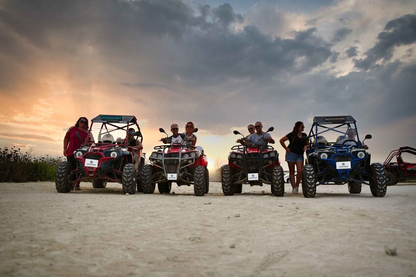 A group of people standing beside ATVs on a sandy surface with a sunset in the background. 2