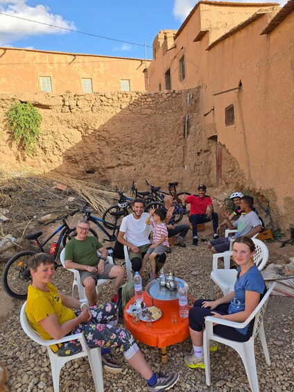 A group of people sitting outdoors with bicycles, a table with food, and a building in the background. 2
