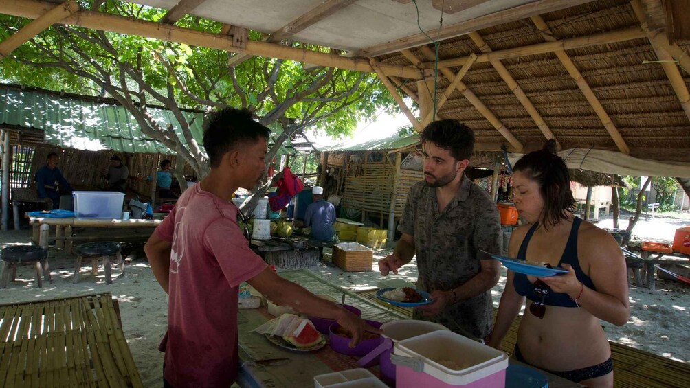 A group of people outdoors under a thatched roof, with tables and food items visible. 2