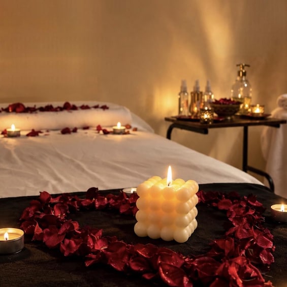 A spa setting with a bed, candles, rose petals, and a table with bottles and a bowl. 1
