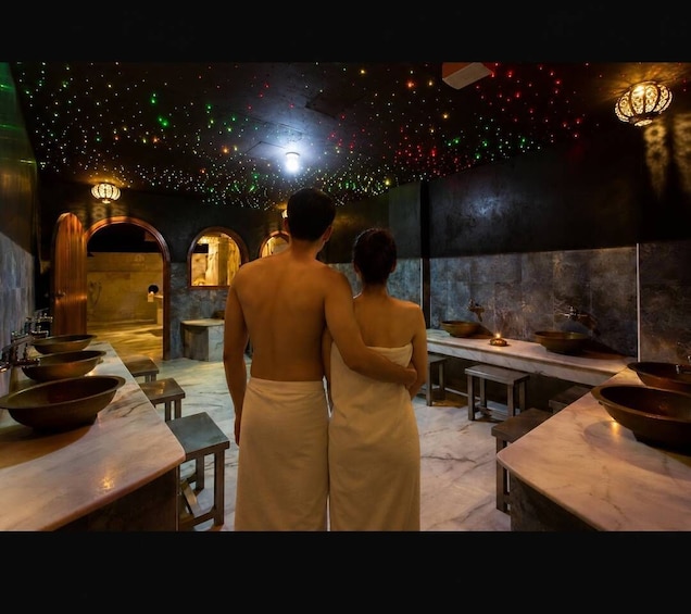 A couple standing in a dimly lit room with a starry ceiling, wooden furniture, and copper bowls on marble tables. 1