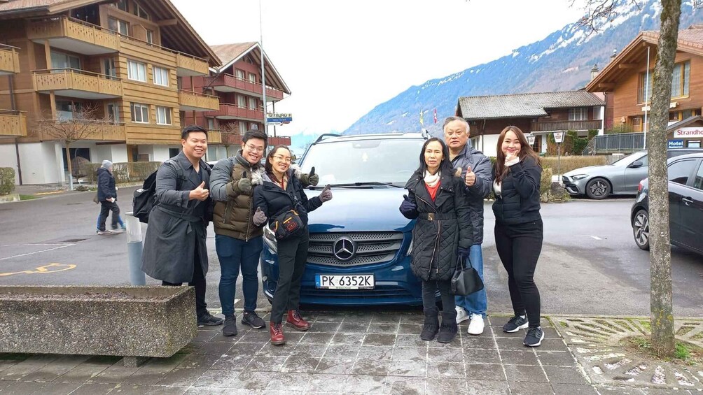 A group of people posing in front of a blue car with a mountain backdrop. 4