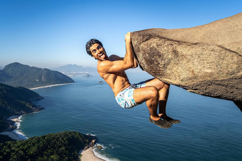 A person climbing a large rock by a coastal area with mountains and a beach in the background. 1