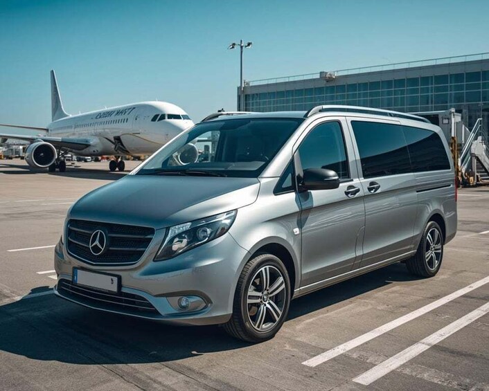 A silver Mercedes-Benz Sprinter van parked on an airport tarmac with an airplane in the background. 1