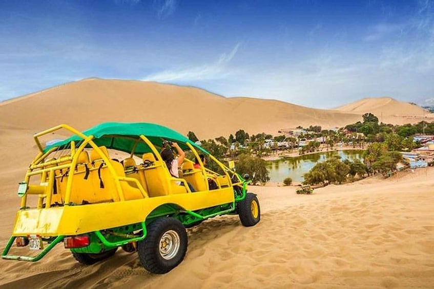A yellow off-road vehicle with a green canopy parked on sand dunes, with a small settlement and water body in the background. 1