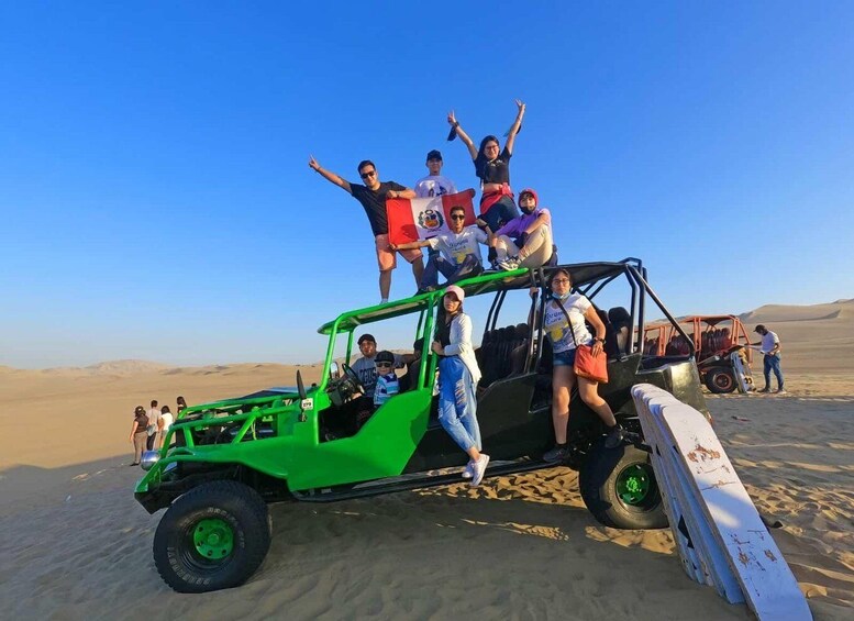 A group of people on a green off-road vehicle in a desert with sand dunes in the background. 5