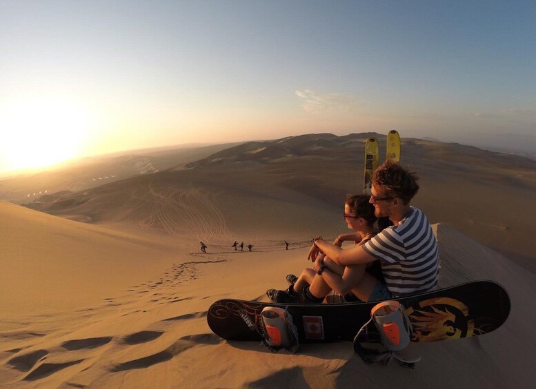 Two people sitting on sand dunes with snowboards and skis nearby. 3