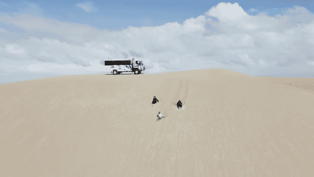 A white truck on a sand dune with three people running towards it. 1