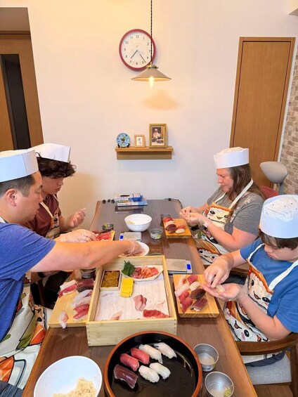 A group of people wearing chef hats, preparing sushi at a wooden table with a clock and framed pictures on the wall. 3