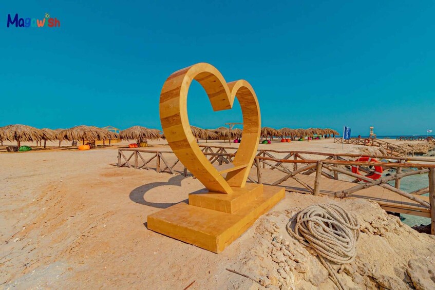 A wooden heart-shaped sculpture on a sandy beach with thatched umbrellas and a wooden pier. 1