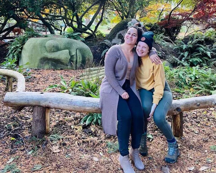 Two people sitting on a log in a forested area with a large rock sculpture in the background. 5