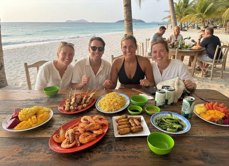 A group of people dining at a wooden table on a beach with a view of the ocean and palm trees. 2