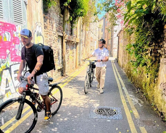 Two cyclists on a narrow urban path with graffiti and greenery. 1