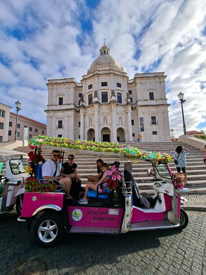A pink and white trolley with passengers and floral decorations, parked in front of a historic building with a dome and columns. 5