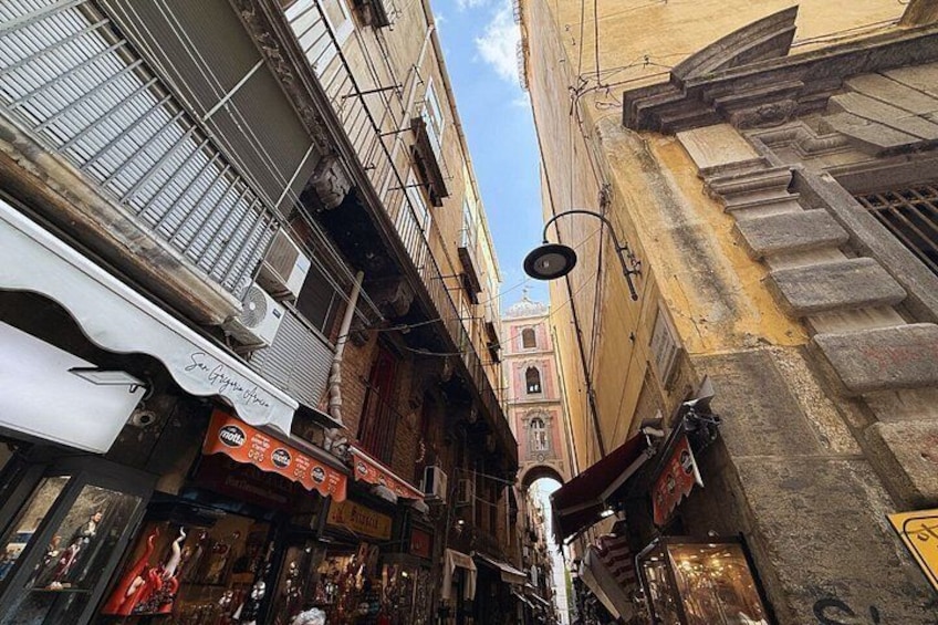 A narrow street with buildings on either side, a street lamp, and a tower in the distance. 2
