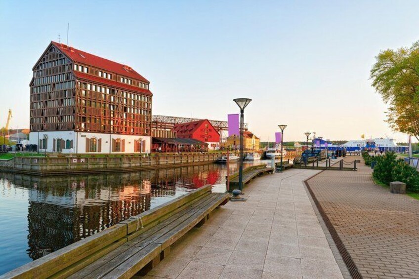 A riverside walkway with a wooden bench, a red-roofed building, and a bridge. 2