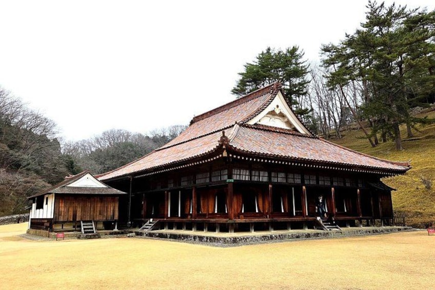 A traditional wooden building with a tiled roof, surrounded by a grassy area and trees. 1