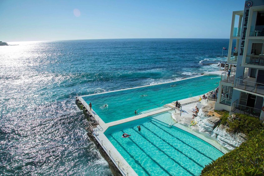 A pool adjacent to a beach with clear blue water, surrounded by rocky cliffs and a building with a sign reading 'Surf Life Saving Australia'. 4