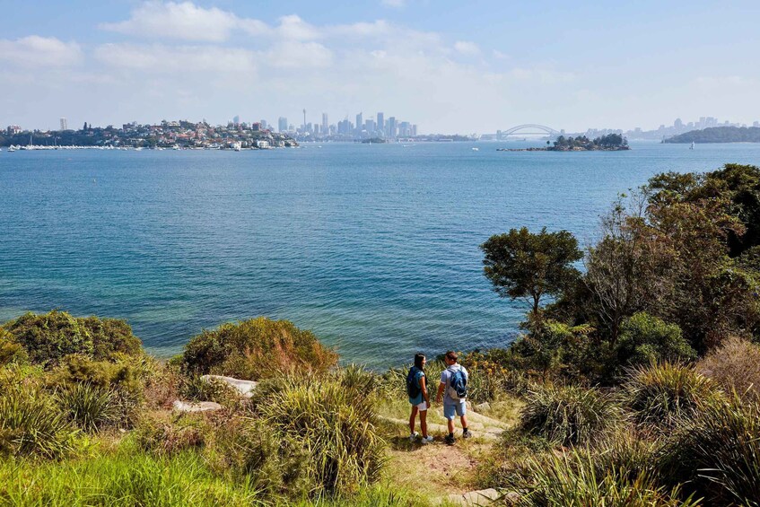 A coastal cityscape with a bridge, two people on a hill overlooking the water, and dense greenery. 2