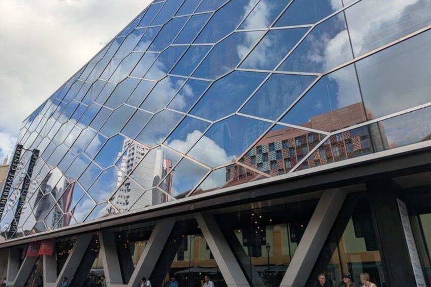 Ein modernes Glasgebäude mit geometrischer Fassade, umgeben von städtischer Bebauung und blauem Himmel. 5