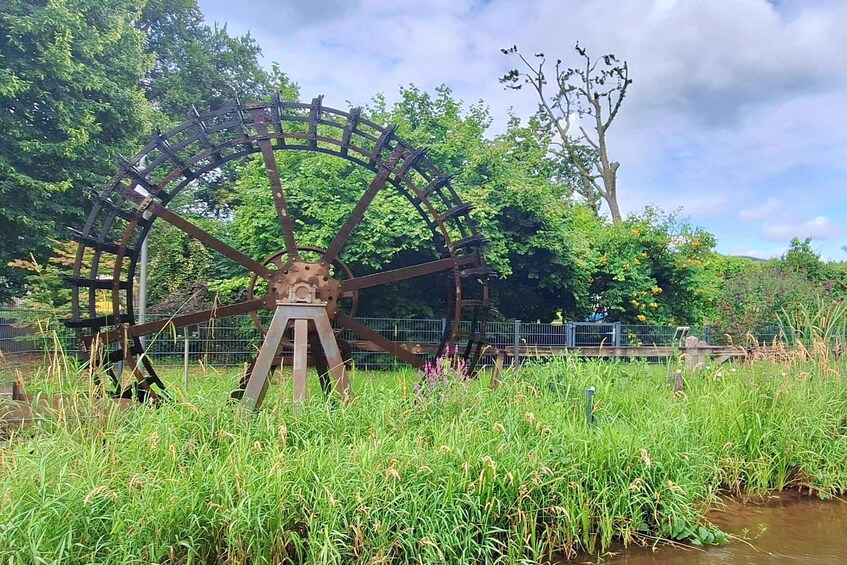 A large water wheel in a grassy area with trees and a fence in the background. 2