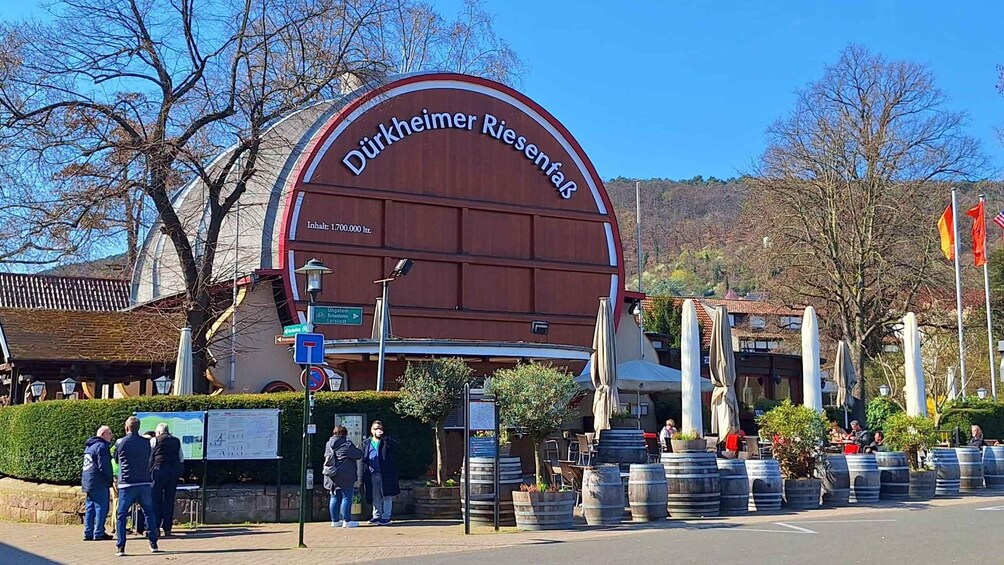 A large sign for 'Dürkheimer Riesentaub' with a capacity of 1,700,000 liters. There are people standing near the sign, and barrels are arranged in front. 1