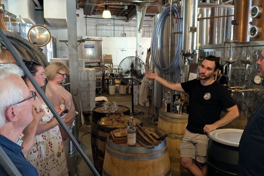 A brewery interior with stainless steel tanks, wooden barrels, and a person gesturing to the audience. 5
