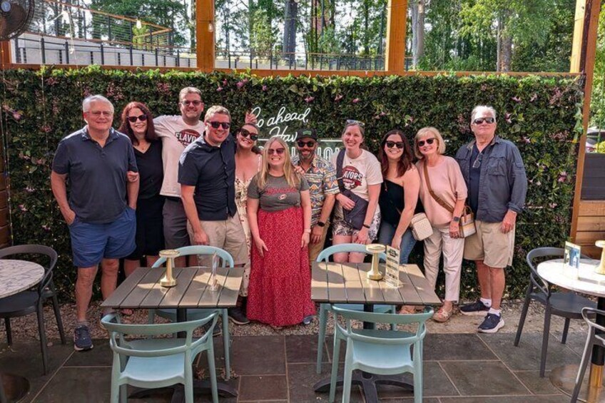 A group of people posing in an outdoor patio area with tables and chairs, surrounded by greenery and a wooden pergola. 1