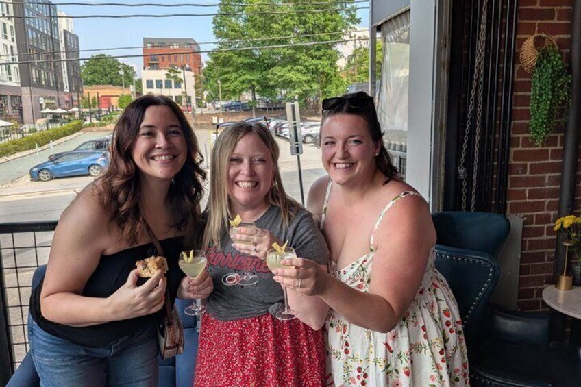 Three people sitting on a balcony with drinks, overlooking a street with buildings and cars. 3